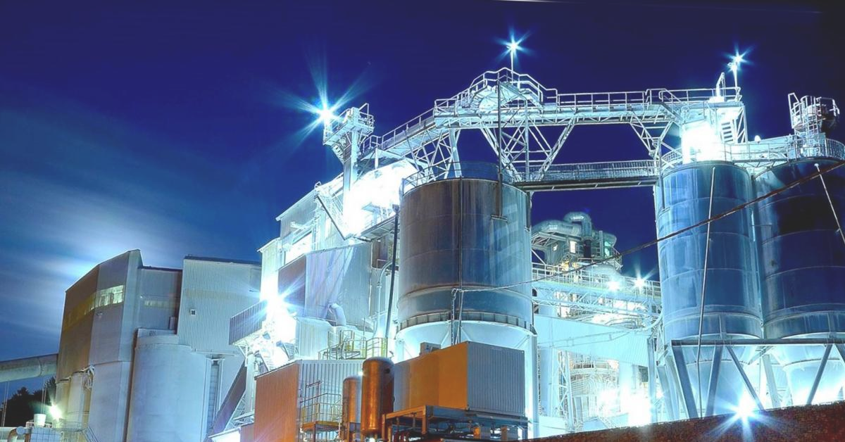 An industrial complex with multiple silos, buildings, and connecting structures, brightly lit against a dark blue night sky.
