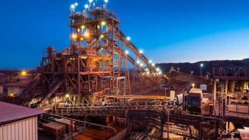 Large mining facility with illuminated processing towers and conveyor belts against a dark blue sky.