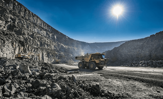 A large yellow dump truck drives through an open-pit mine with excavators and rocky walls under a bright sun.
