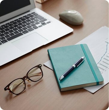 Laptop, mouse, glasses, teal notebook with a pen, and papers with a graph on a wooden desk.