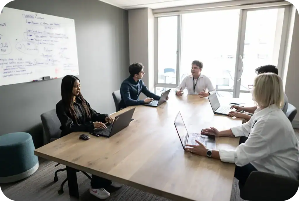 Five people in a meeting room with laptops; one talks, three type, one listens.