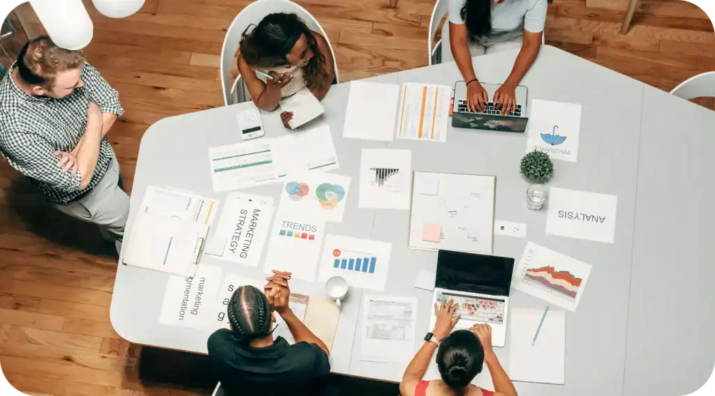 Overhead view of five people around a table with laptops, charts, and documents during a meeting.